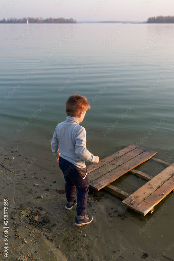 Little kid boy on a wooden pier on a lake, a river looks into the distance at the horizon
