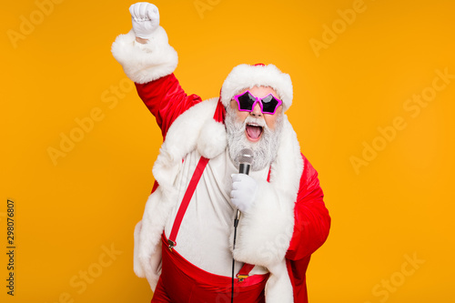 Grey haired stylish christmas grandfather in red hat cap celebrate x-mas party hold microphone sing noel carols feel funky with big belly wear suspenders isolated over yellow color background