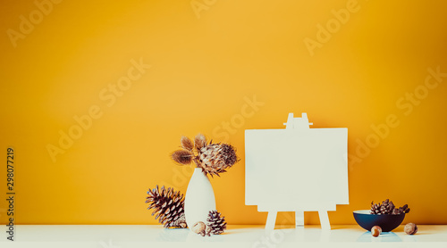 Εκτύπωση καμβά Composition of dried flowers, thorns in light vase, pine cones and blank canvas on a stand on ocher color wall background