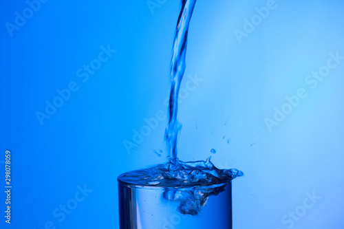 Chorro de agua llenando y rebosando un vaso de agua de cristal o plástico transparente sobre fondo azul