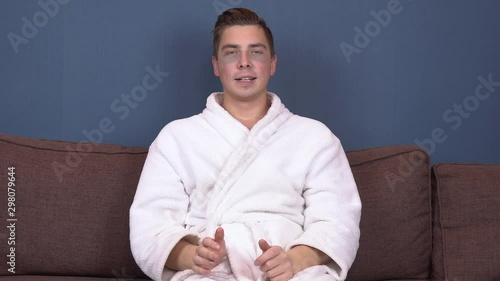 A young man with patches on his face relaxes. Spa treatments. Blue hydrogel patches for facial rejuvenation. A man sits in a white coat on the couch. photo