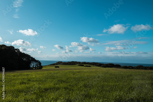 Campo verde despejado con cielo azulado y nubes y mar de fondo.