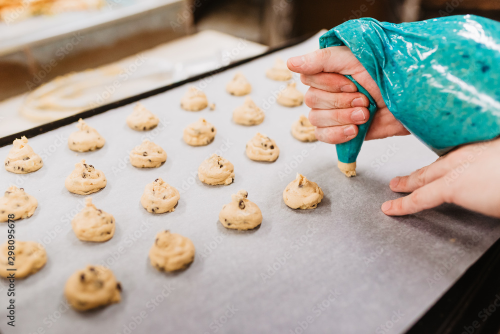 Anonymous cook squeezing fresh pastry dough on tray with paper while working on blurred background of bakery Anonymous cook squeezing fresh pastry dough on tray with paper while working on blurred background of bakery