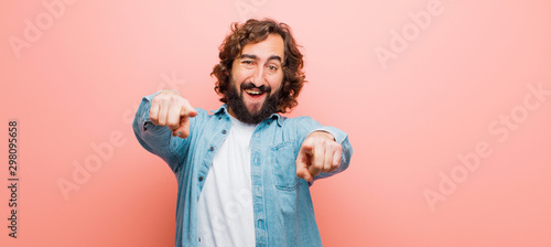 young bearded crazy man feeling happy and confident, pointing to camera with both hands and laughing, choosing you against flat color wall