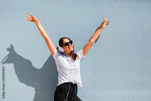 Carefree beautiful woman in casual outfit and headphones on neck standing with hands up beside blue wall of building on city street