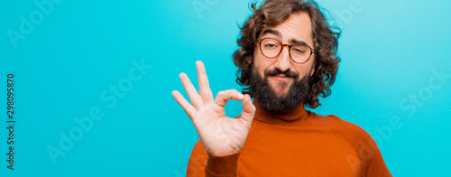 young bearded crazy man feeling happy, relaxed and satisfied, showing approval with okay gesture, smiling against flat color wall