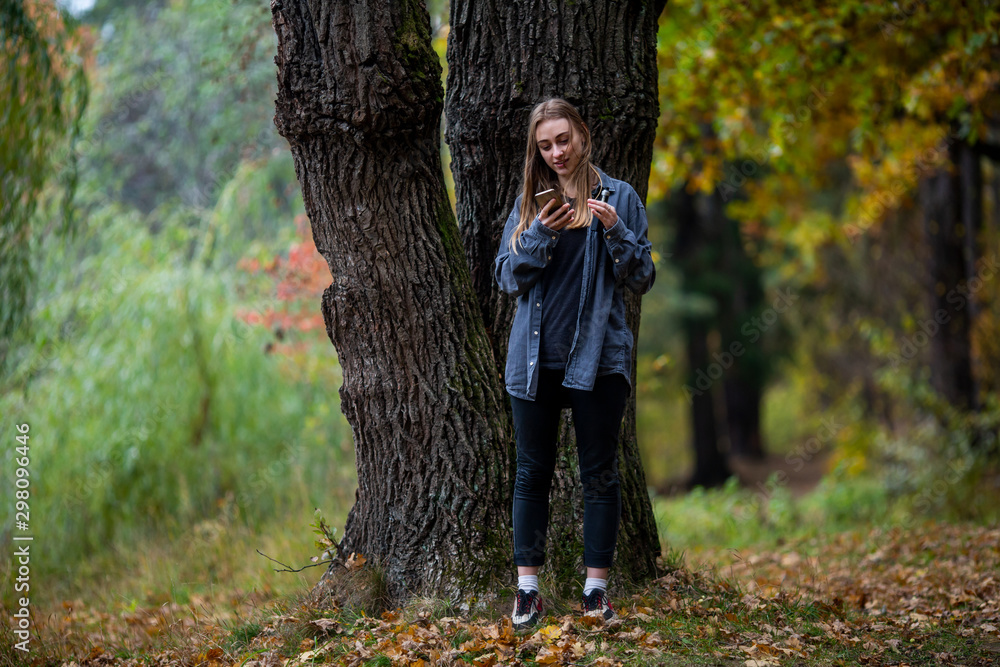 Teenager with phone and vape. Young beautiful white teenage girl in casual clothes and with smartphone on the street in the dark forest in the autumn evening. Teenager with phone and vape. Young beautiful white teenage girl in casual clothes and with smartphone on the street in the dark forest in the autumn evening.