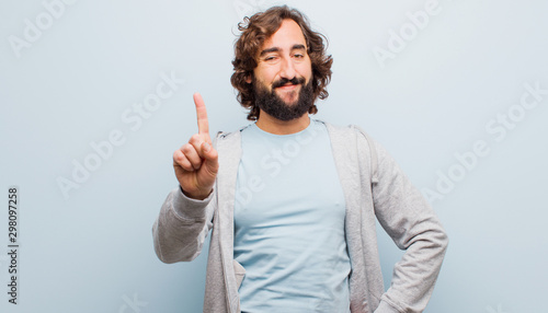 young bearded crazy man smiling and looking friendly, showing number one or first with hand forward, counting down against flat color wall
