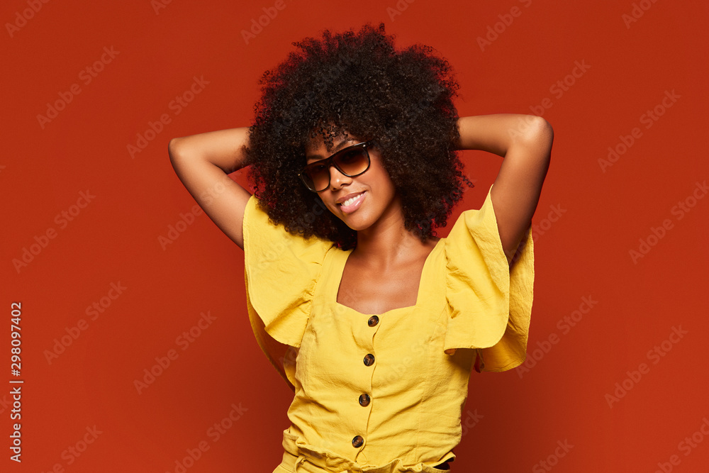Modern young black woman with Afro hairstyle holding hands behind head while smiling at camera on bright red background Modern young black woman with Afro hairstyle holding hands behind head while smiling at camera on bright red background