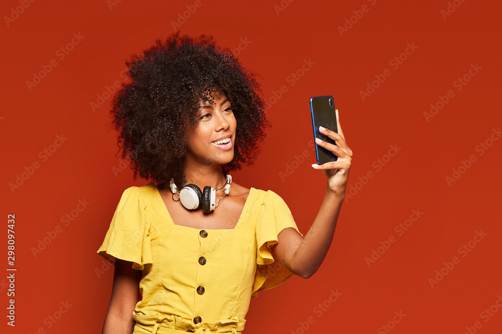 Modern happy ethnic woman with curly Afro hair wearing headphones on neck and taking selfie with mobile phone on vivid red backdrop Modern happy ethnic woman with curly Afro hair wearing headphones on neck and taking selfie with mobile phone on vivid red backdrop