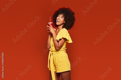 Cheerful young African American lady with curly hair holding red jar with straw and enjoying beverage on red background looking at camera photo