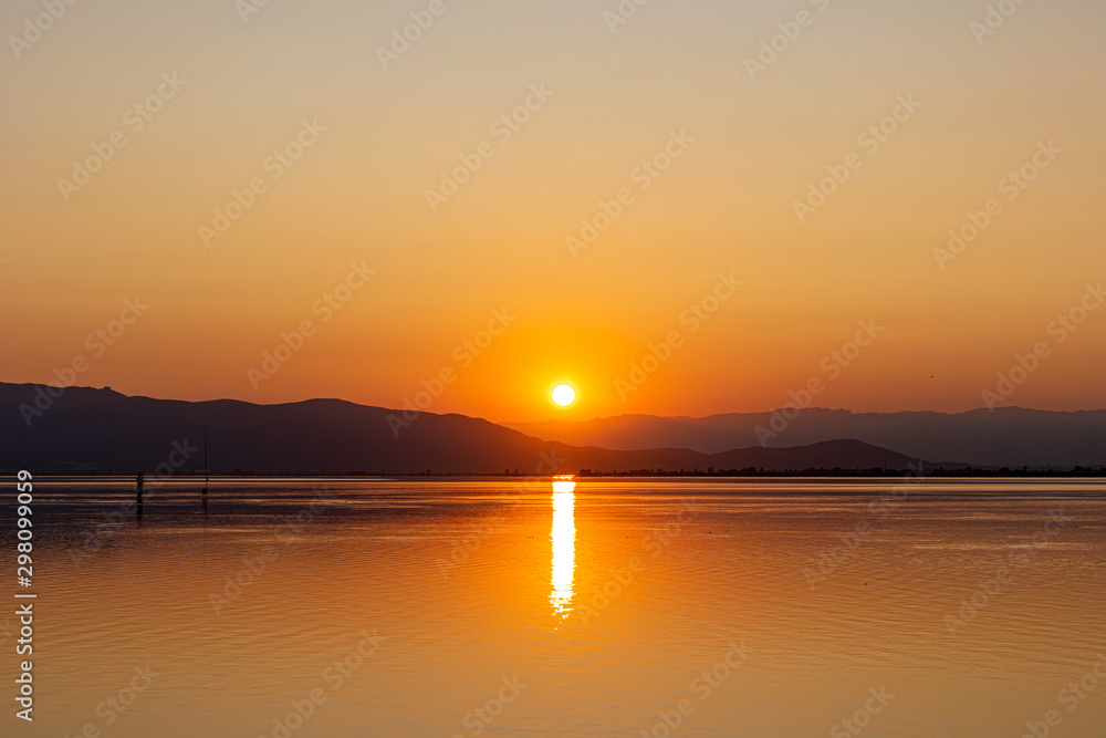 Orange sun going down behind dark hills reflecting in peaceful slightly rippled water creating romantic seascape