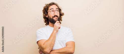 young bearded crazy man feeling thoughtful, wondering or imagining ideas, daydreaming and looking up to copy space against flat color wall photo