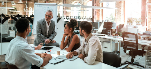 Working process. Mature businessman talking about something with his young colleagues while sitting together at the office table in the modern office