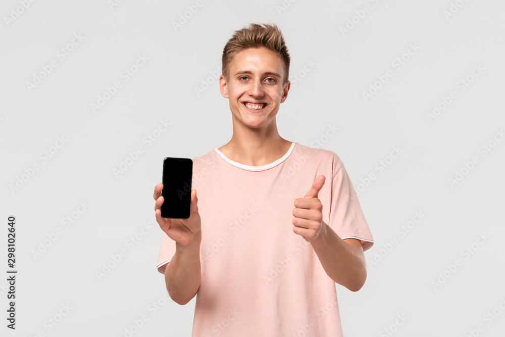 Smiling blond teenager in a pink tee pointing at the screen of a smartphone isolated over light grey background. Smiling blond teenager in a pink tee pointing at the screen of a smartphone isolated over light grey background.