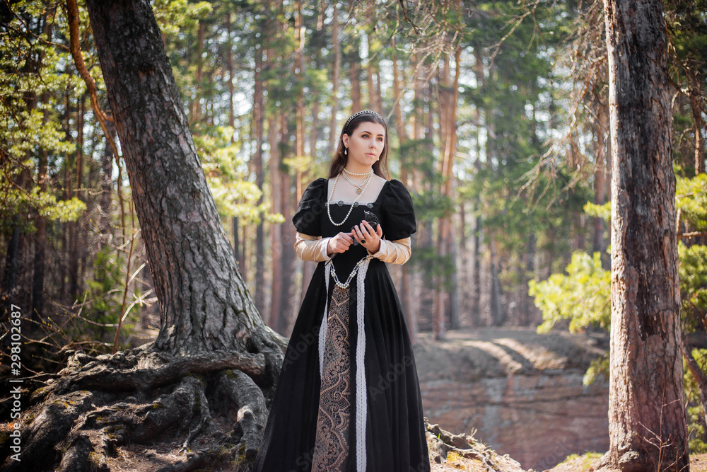 Fototapeta premium Young, beautiful girl in a black medieval dress with a steel rose in hand, walking through the forest.