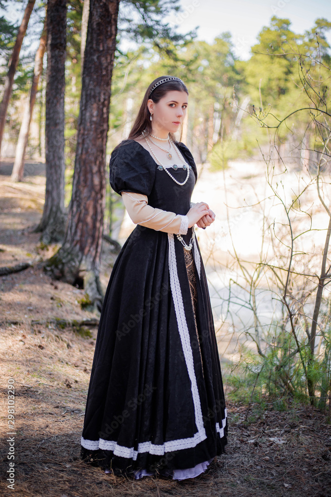 Fototapeta premium Young, beautiful girl in a black medieval dress walks through the gloomy pine forest.