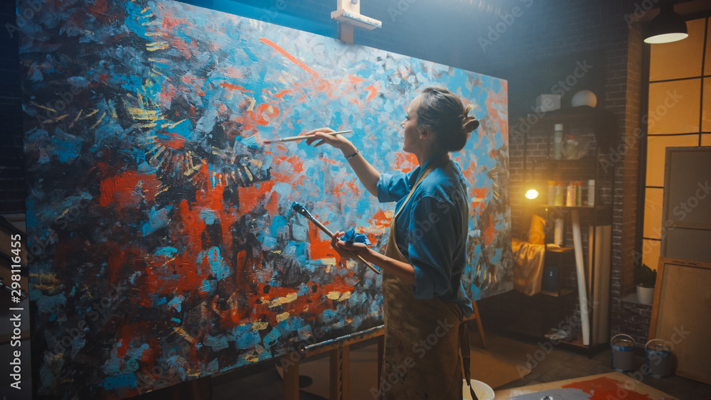 Talented Female Artist Works on Abstract Oil Painting, Using Paint Brush She Creates Modern Masterpiece. Dark and Messy Creative Studio where Large Canvas Stands on Easel Illuminated.