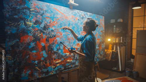 Talented Female Artist Works on Abstract Oil Painting, Using Paint Brush She Creates Modern Masterpiece. Dark and Messy Creative Studio where Large Canvas Stands on Easel Illuminated. photo