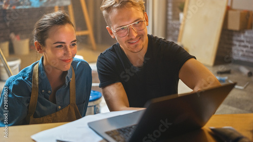 Obraz na plátně Couple of Artists Sitting in their Studio Use Laptop Computer, Working on a Project, Talking and Smiling