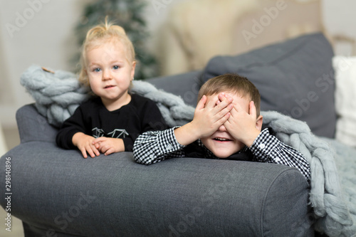 Little girl and boy lying t on the couch covered with a gray knitted blanket and smiling. Brother and sister grimacing and fooling around on the background of New Year's decor and Christmas tree