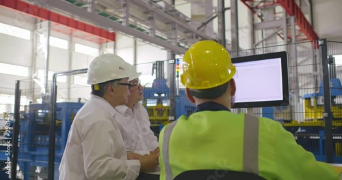 Two engineers white collars discussing with workman in uniform data at display of computer of CNC machine operation. Three colleagues have conversation at industrial background
