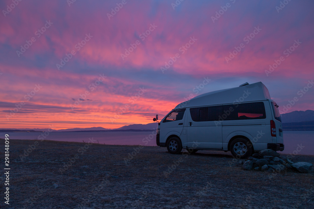 A campervan is standing at Lake Tekapo, New Zealand, looking into the sunset. The sky is scattered with some clouds with vibrant red, purple and orange colors.