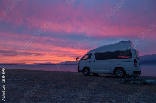 A campervan is standing at Lake Tekapo, New Zealand, looking into the sunset. The sky is scattered with some clouds with vibrant red, purple and orange colors. © djr-photography