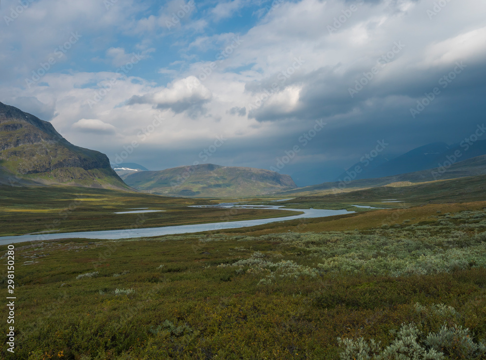Beautiful wild Lapland nature landscape with blue glacial river, birch tree bushes, snow capped mountains and dramatic clouds. Northern Sweden summer at Kungsleden hiking trail.