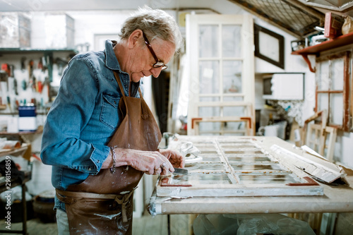 Side view of senior male owner scraping window frame on workbench at store workshop photo