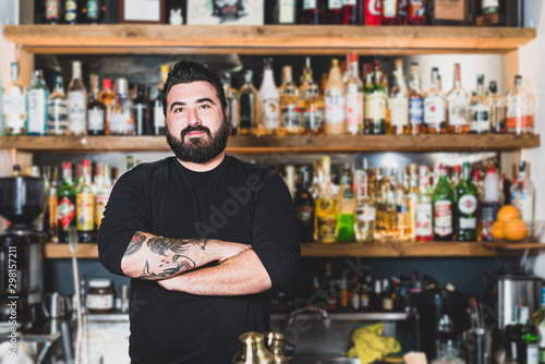 Young hipster bartender with thick beard smiles at the counter with a background of spirits behind him