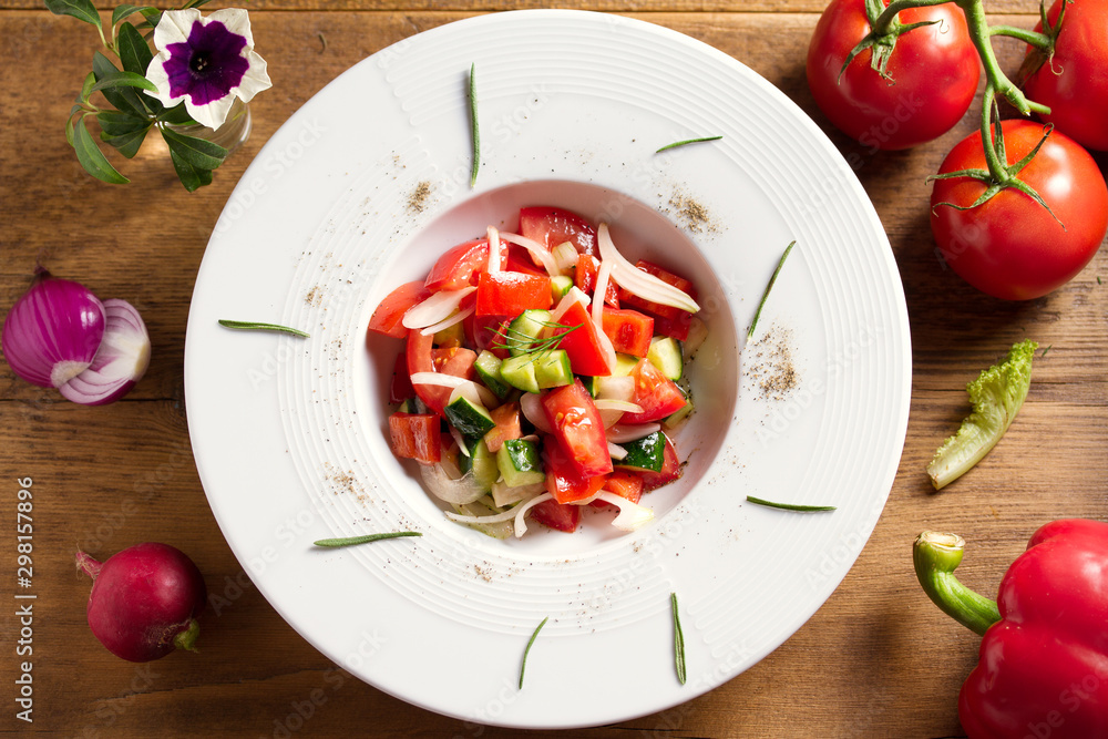 Vegetable salad of fresh tomatoes, cucumbers, bell pepper, onions and herbs in white bowl, wooden table. Overhead, horizontal image