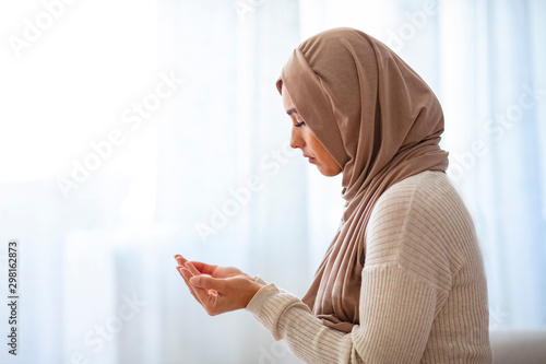 Muslim woman in beige hijab and traditional clothes praying for Allah, copy space. Muslim woman with hijab praying indoor at bright window. Young Muslim woman Praying