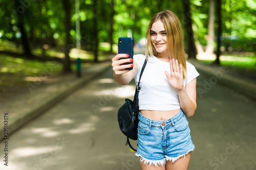 Beauty teenage girl taking a selfie on smartphone outdoors in park on sunny day.