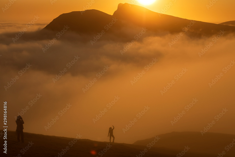  Clouds covered the mountain. The sun painted the sky and the mount in golden color. Silhouettes of girls on a background of mounts and sun, Faroe Islands. Soft focus. 