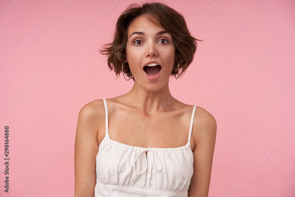 Surprised pretty young brunette woman with short haircut looking at camera with wide eyes and mouth opened, posing over pink background with hands down - obrazy, fototapety, plakaty 