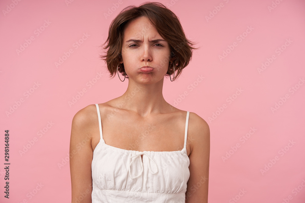 Offended brunette woman with casual hairstyle pursing lips and looking at camera with squint, wearing white top while standing over pink background with hands down Offended brunette woman with casual hairstyle pursing lips and looking at camera with squint, wearing white top while standing over pink background with hands down