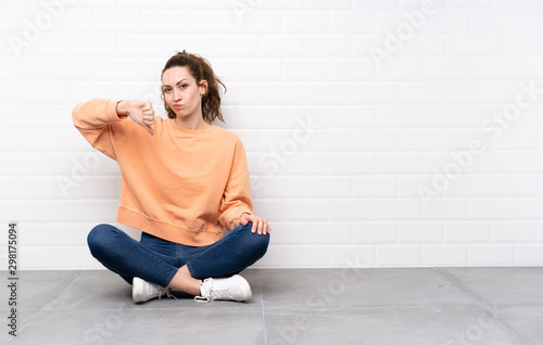 Young woman with curly hair sitting on the floor showing thumb down sign