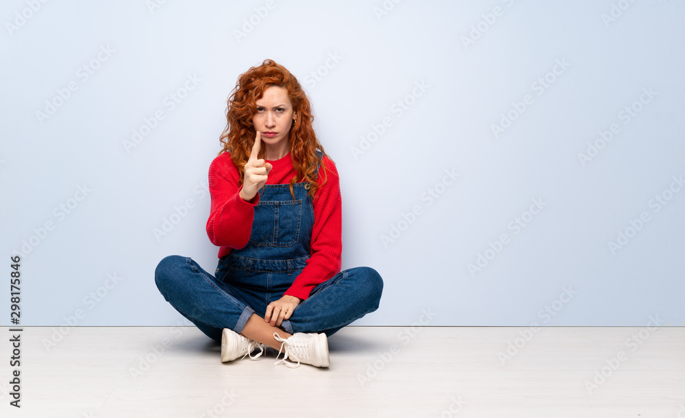 Redhead woman with overalls sitting on the floor frustrated and pointing to the front