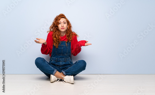 Redhead woman with overalls sitting on the floor having doubts while raising hands