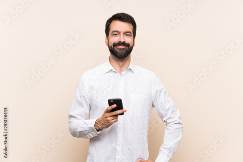 Young man with beard holding a mobile posing with arms at hip and smiling