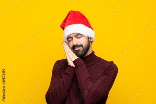 Man with christmas hat over isolated yellow background making sleep gesture in dorable expression
