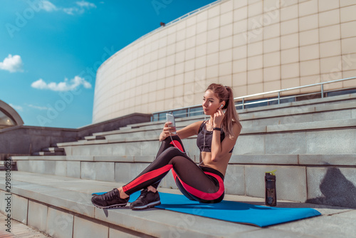Girl athlete sits mat yoga gymnastics, phone listens music headphones, rest workout, summer day city. Enjoys listens podcast lecture on Internet. Active lifestyle fitness, fashionable stylish woman.
