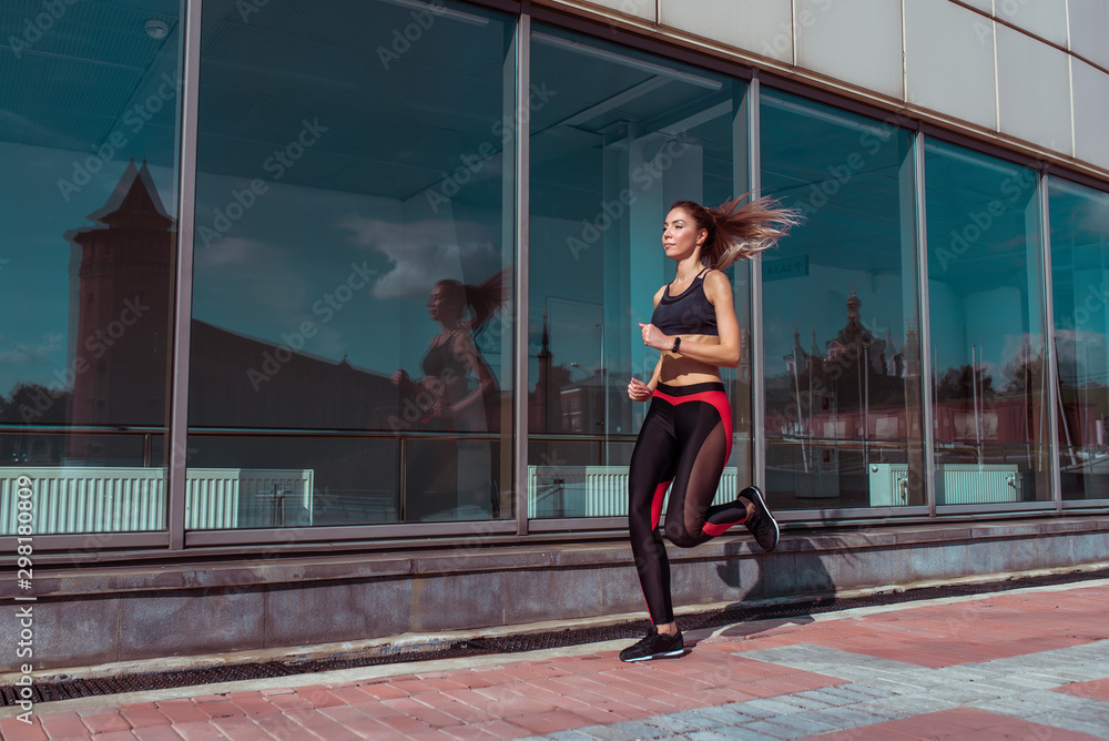 Athletics training, day morning. Young athletic woman running in summer on jogging in city, background glass windows of building, sportswear. Active fitness lifestyle, free space text motivation.