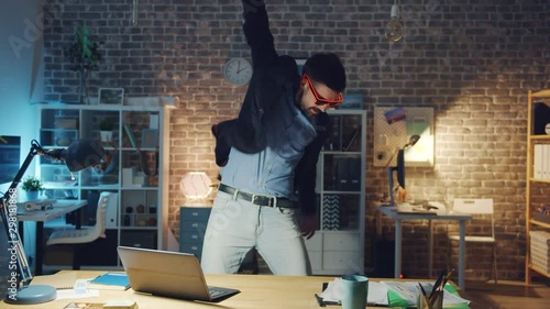 Handsome office worker in stylish glasses is dancing in office room at night, loft style workplace is visible in background. People, joy and modern lifestyle concept. photo