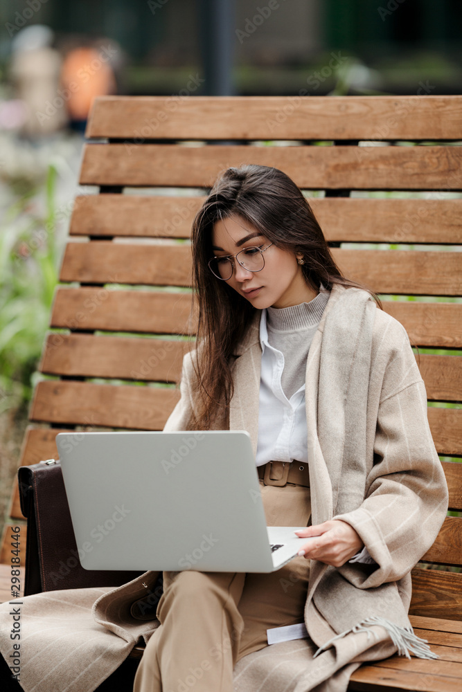 A beautiful businesswoman of Asian appearance looks through working documents on her laptop sitting in an autumn Park. Vertical photo A beautiful businesswoman of Asian appearance looks through working documents on her laptop sitting in an autumn Park. Vertical photo