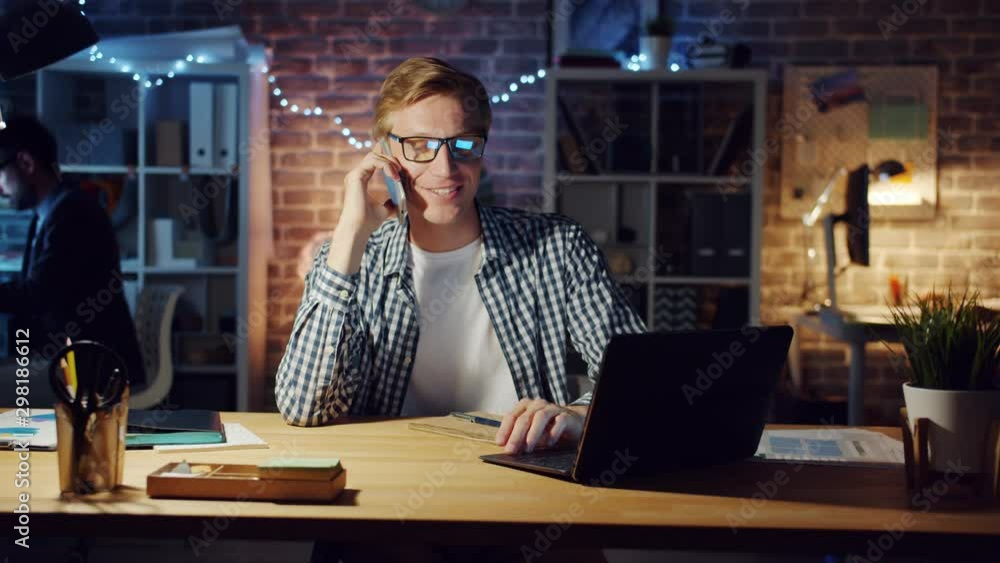 Cheerful office worker speaking on mobile phone and working with laptop in dark office at night sitting at table smiling. Happy people, communication and business concept. Cheerful office worker speaking on mobile phone and working with laptop in dark office at night sitting at table smiling. Happy people, communication and business concept.