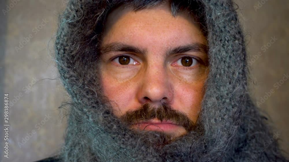 A man in a warm shawl on his head. A bearded man has a sick look. Blinks eyes. Against the background of gray wallpaper on the wall. A man in a warm shawl on his head. A bearded man has a sick look. Blinks eyes. Against the background of gray wallpaper on the wall.