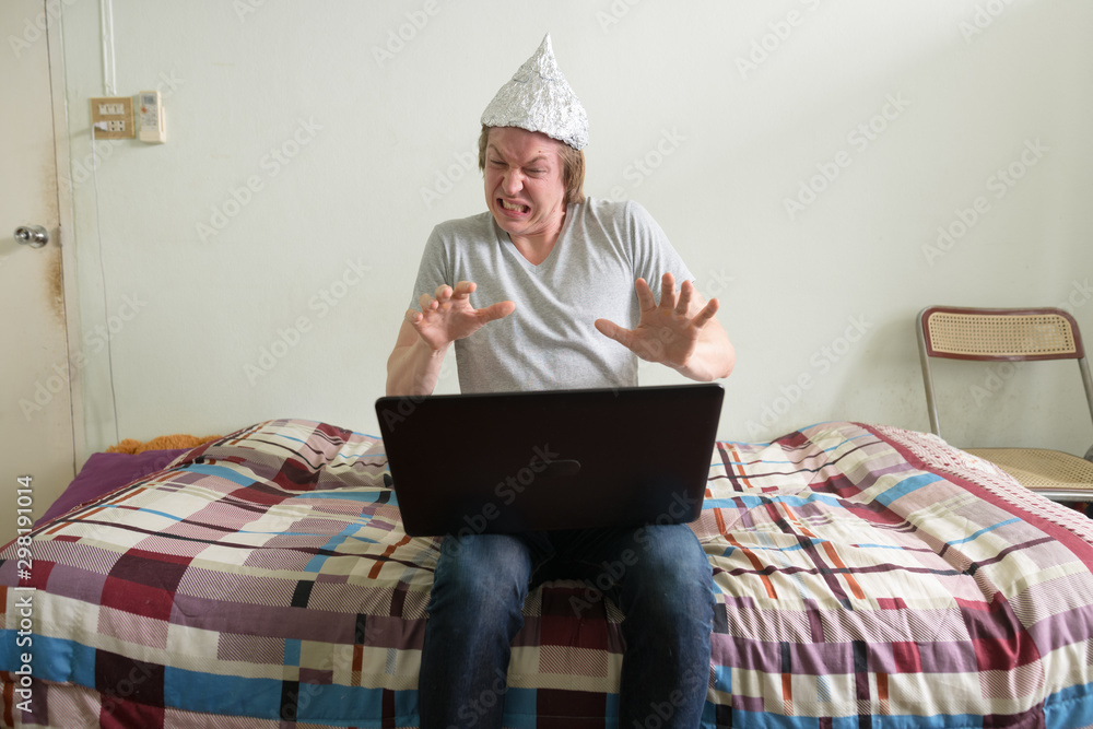 Stressed young man with tin foil hat using laptop and showing stop gesture in the bedroom Stressed young man with tin foil hat using laptop and showing stop gesture in the bedroom