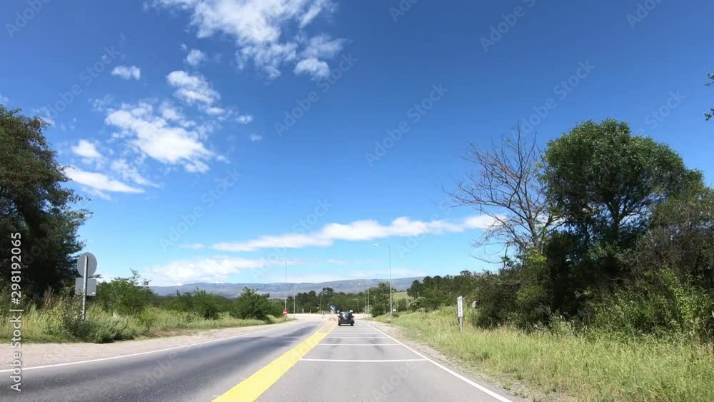 Driving on a road through grasslands, rocky hills and woody landscape. Taking roundabout. Background of mountains. Traveling along native ecosystems. Cordoba, Argentina Driving on a road through grasslands, rocky hills and woody landscape. Taking roundabout. Background of mountains. Traveling along native ecosystems. Cordoba, Argentina
